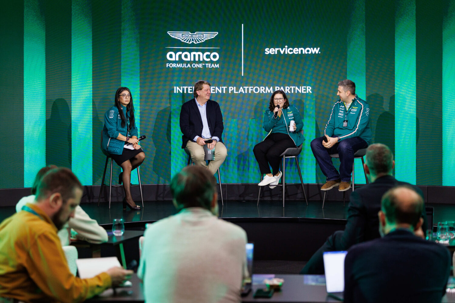 Vier panelleden zitten op krukjes op het podium tijdens een discussie, met publiek op de voorgrond en "aramco Formula One Team" op de achtergrond.