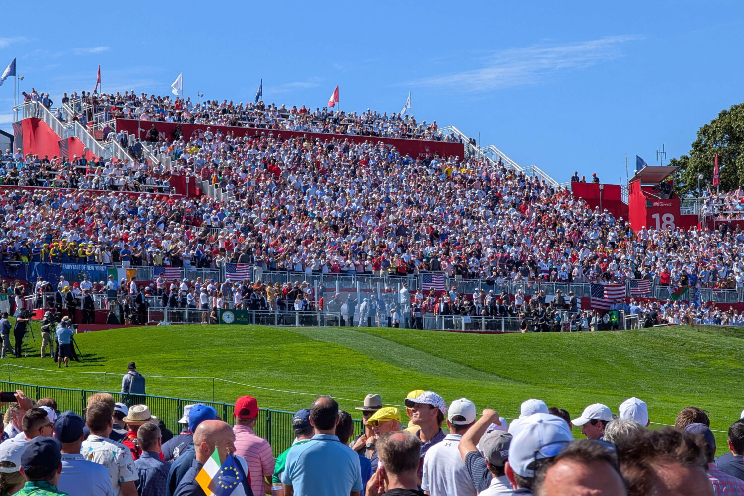 Spectators_Ryder_Cup Een grote menigte vult tribunes in stadionstijl met uitzicht op een golfbaan onder een strakblauwe hemel, met mensen verzameld op het gras en Amerikaanse vlaggen zichtbaar.