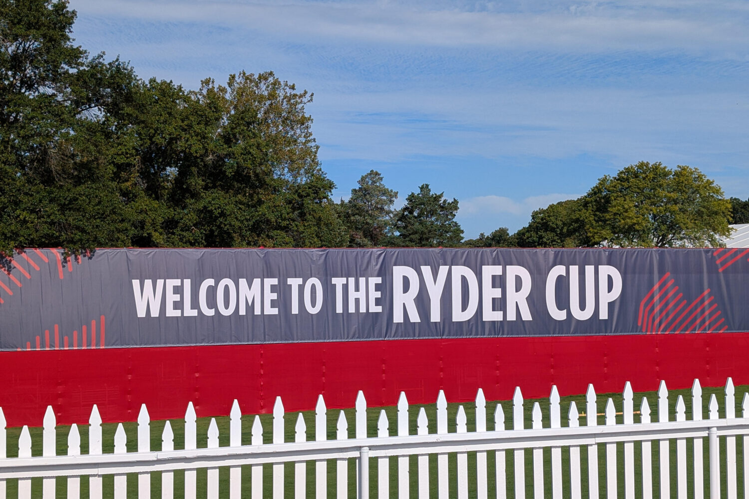 Welcome_Ryder_Cup Een groot bord met de tekst "Welcome to the Ryder Cup" staat achter een wit hek, met bomen en een blauwe lucht op de achtergrond.