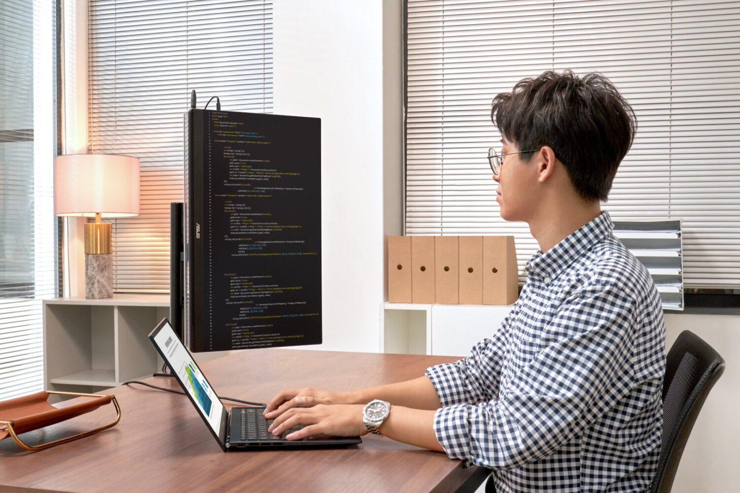 Een persoon met bril en geruit overhemd typt op een laptop aan een bureau met een verticale monitor die code weergeeft, in een moderne kantooromgeving.