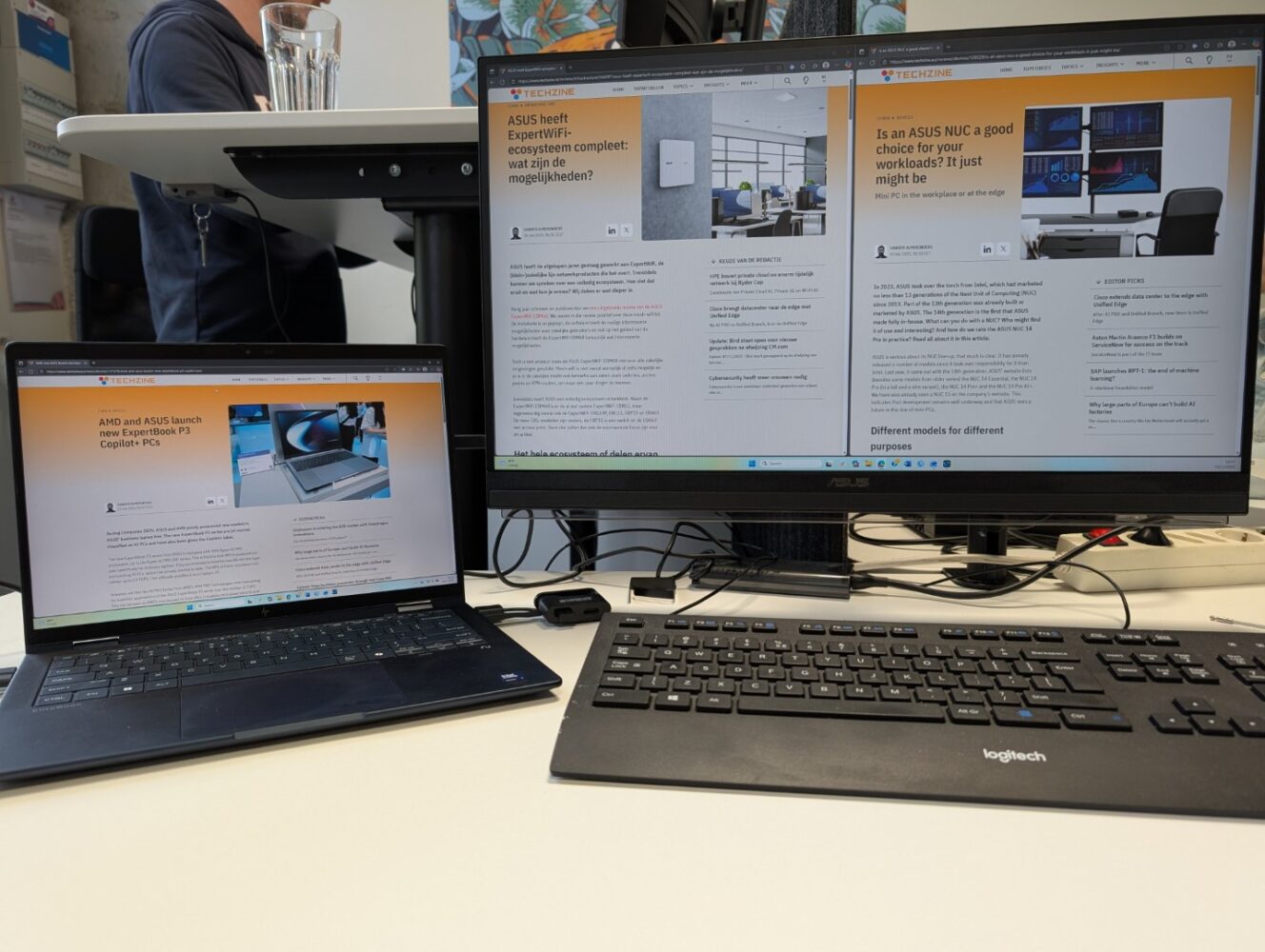 Een laptop en een monitor op een bureau tonen soortgelijke webpagina's over technologische onderwerpen; een toetsenbord en muis bevinden zich voor de monitor.