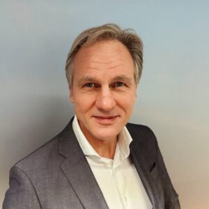 Een man van middelbare leeftijd met grijs haar in een donker colbert en wit overhemd staat tegen een lichtblauwe achtergrond, kijkt naar de camera en glimlacht lichtjes.