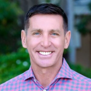 Een man met kort bruin haar en een rood en blauw geruit overhemd lacht naar de camera, met groen op de achtergrond.