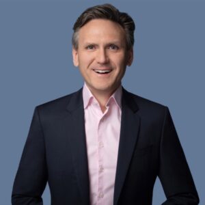 Een man in een donker pak en lichtroze overhemd staat te glimlachen tegen een effen blauwe achtergrond.