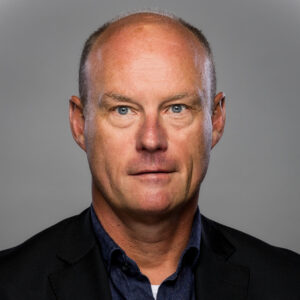 Een man van middelbare leeftijd met een kaal hoofd in een donker colbert en blauw overhemd, kijkt naar de camera met een neutrale uitdrukking tegen een grijze achtergrond.