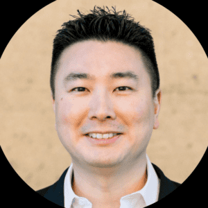 Een man met kort, donker haar draagt een wit overhemd en donkere blazer, glimlachend voor een beige achtergrond.