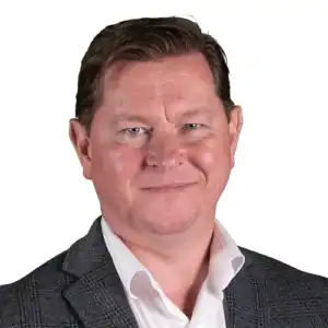 Man met kort bruin haar draagt een wit overhemd en grijze blazer, kijkt naar voren en glimlacht, op een effen witte achtergrond.