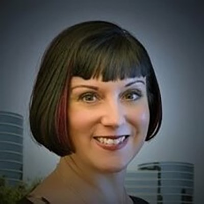 kellyn-potvin-gorman Een vrouw met kort donker haar en pony lacht naar de camera. Op de onscherpe achtergrond zijn drie moderne kantoorgebouwen te zien.