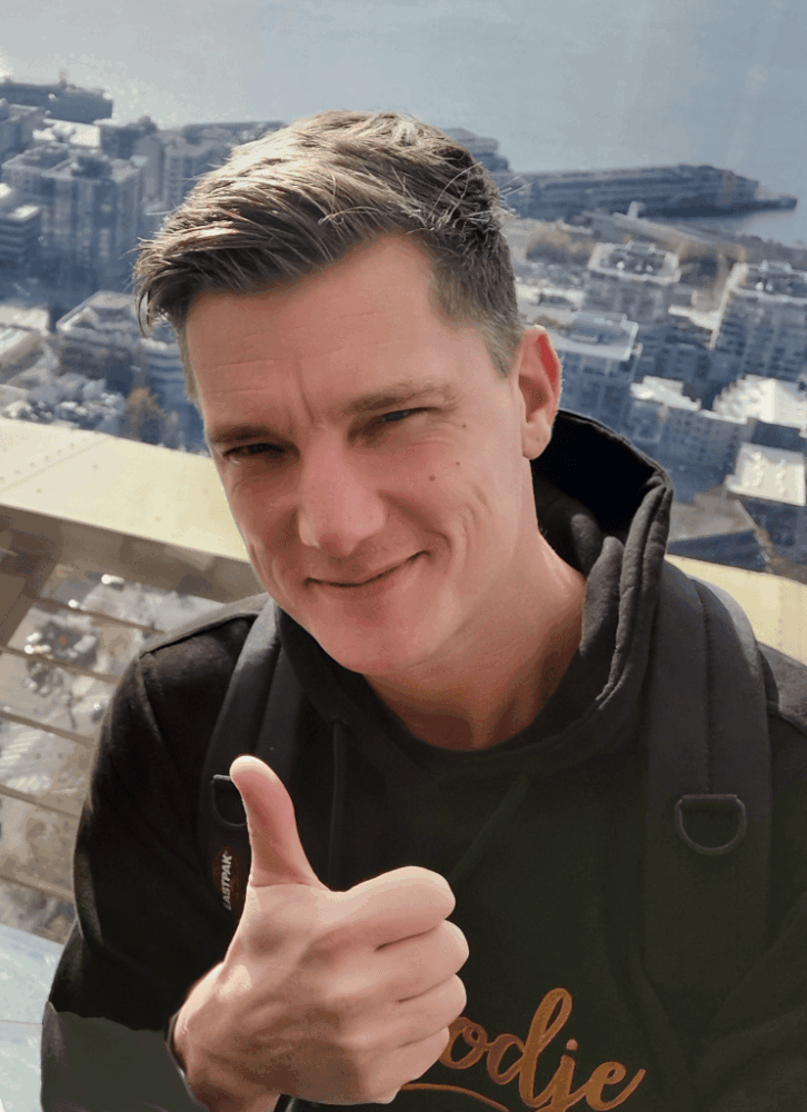 tonie-seattle-2022-deflipped-743×1024 Een persoon met kort bruin haar, een zwarte hoodie en rugzak geeft een duim omhoog met een stadsgezicht en water zichtbaar op de achtergrond.