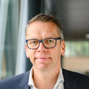 Een man van middelbare leeftijd met kort haar en een blauwe bril, die een donker jasje en wit overhemd draagt, staat binnen bij grote ramen.