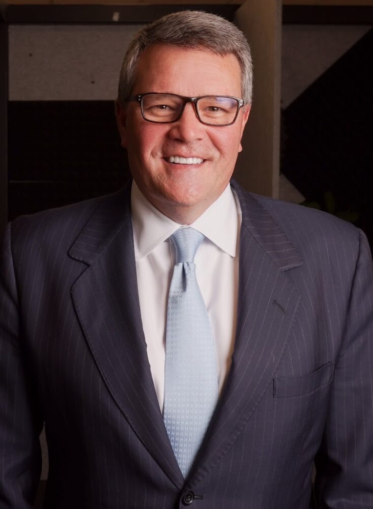 Een man met een bril, een donker krijtstreeppak, een lichtblauwe stropdas en een wit overhemd staat binnen en glimlacht naar de camera.