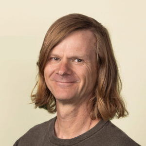 Een persoon met schouderlang lichtbruin haar die een donkere trui met textuur draagt, staat tegen een effen, lichtgekleurde achtergrond en glimlacht naar de camera.