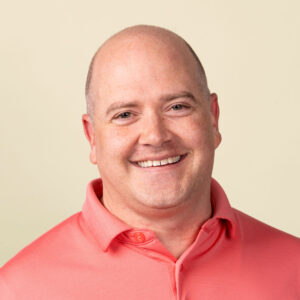 Een man van middelbare leeftijd met een kaalgeschoren hoofd lacht naar de camera terwijl hij een roze overhemd draagt tegen een effen lichte achtergrond.