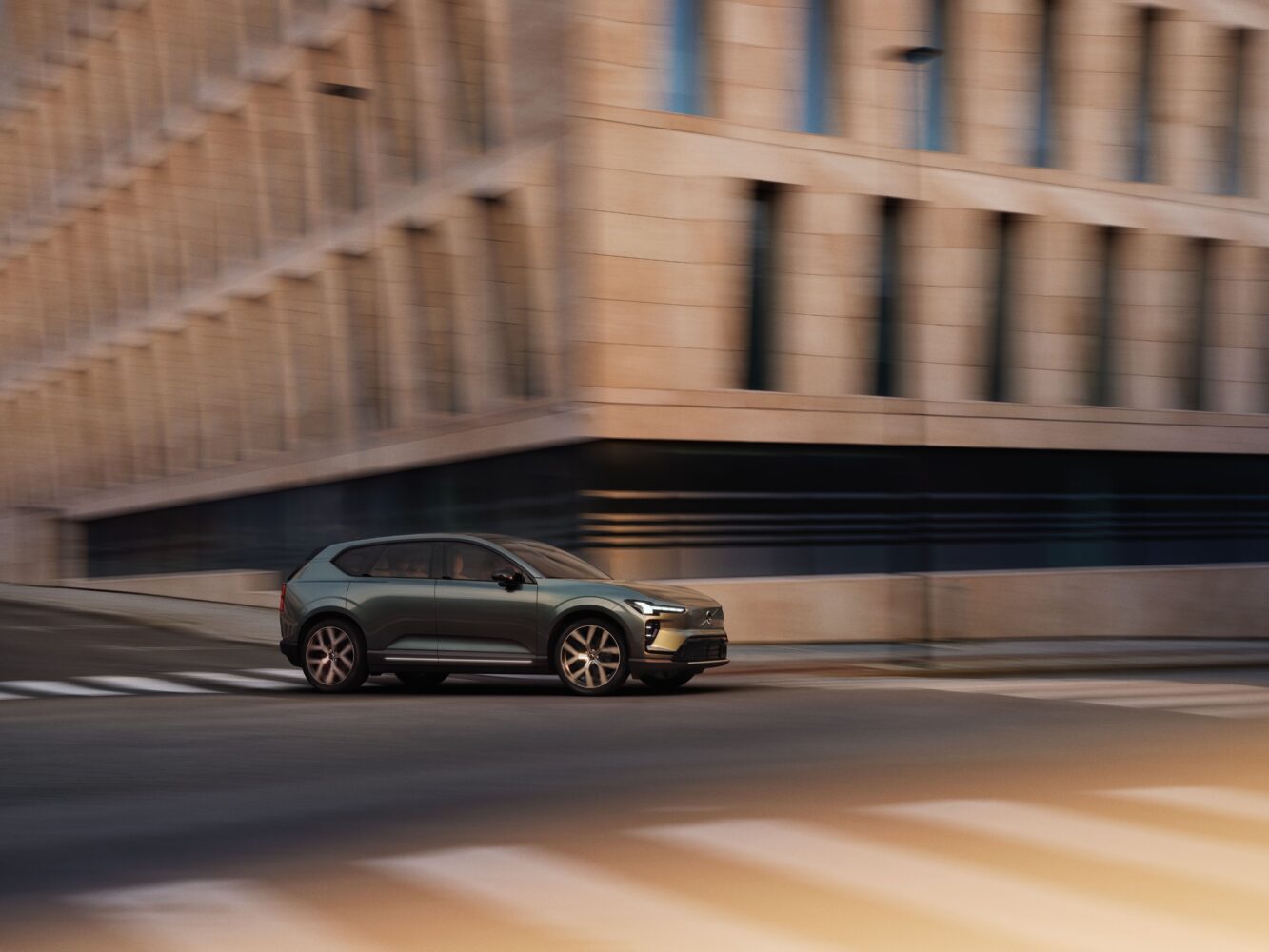 JPG Full-TIM_1157_EX60_3Q_FRONT_DRIVING Een moderne grijze SUV rijdt over een leeg kruispunt in de stad, met een vage achtergrond van een beige kantoorgebouw.