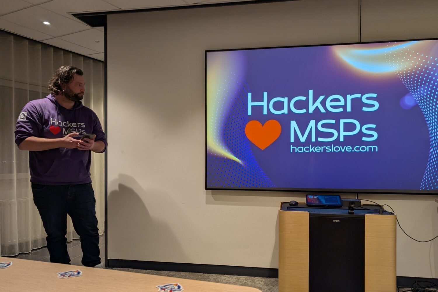 Een persoon staat bij een groot scherm met de tekst "Hackers ♥ MSPs hackerslove.com" in een conferentieruimte, met daarop een Anthropic Claude hack-sessie.