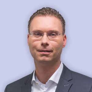 Een man met een bril en een colbert over een wit overhemd kijkt naar voren tegen een effen lichte achtergrond.