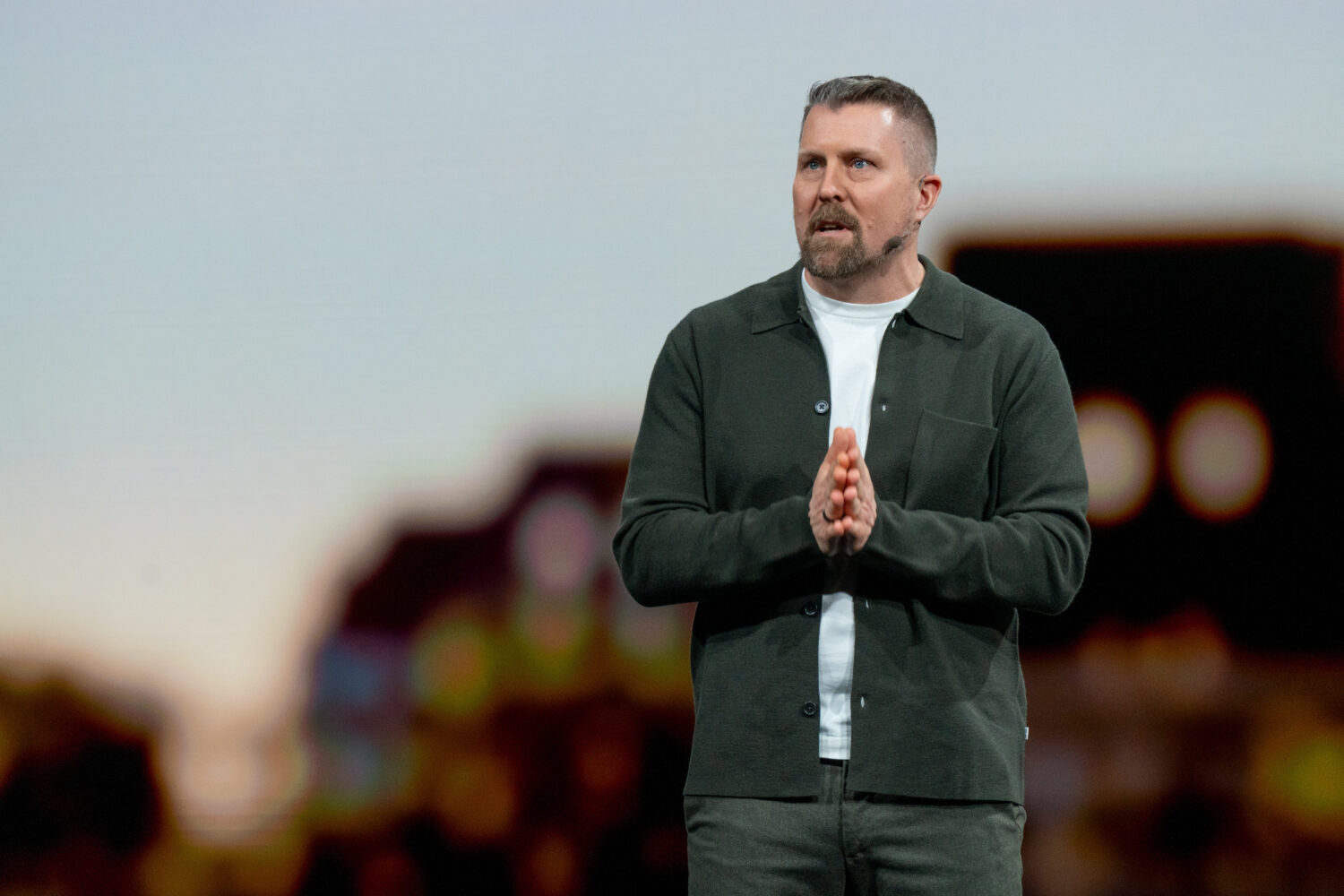 Volvo_EX60_Reveal_event(15) Een man met kort haar en een baard staat op het podium, de handen ineen geklemd, sprekend voor een wazige achtergrond.