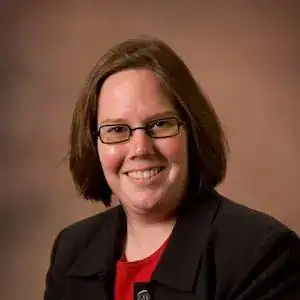 Een vrouw met kort bruin haar en een bril lacht naar de camera, draagt een donkere blazer en een rood topje voor een bruine gradiënt achtergrond.