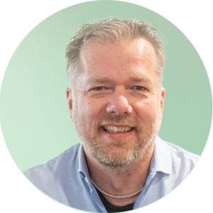 Glimlachende man van middelbare leeftijd met kort, licht haar en een baard, draagt een lichtblauw shirt en poseert tegen een lichtgroene achtergrond.