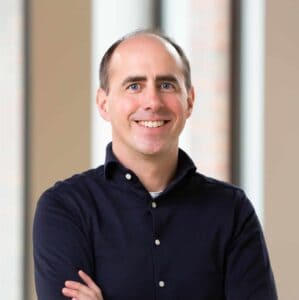 Een man met kort bruin haar en een marineblauw overhemd met knoopsluiting glimlacht met gekruiste armen en staat binnen voor vage ramen.