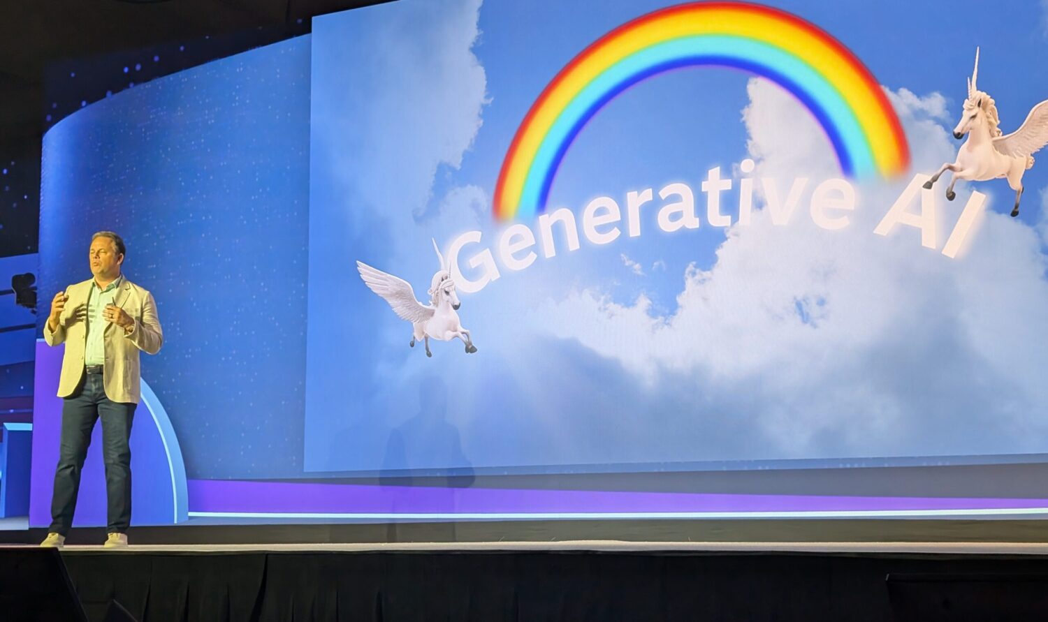 Een persoon staat op het podium naast een groot scherm waarop "Generative AI" wordt weergegeven met een regenboog, hemelachtergrond en twee vliegende eenhoorns.
