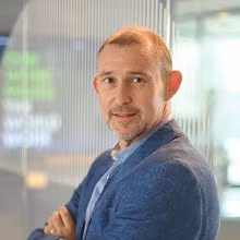 Een man met kort haar en een baard in een blauwe blazer staat binnen met zijn armen over elkaar en kijkt naar de camera.