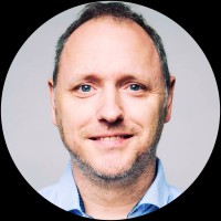 Een man met kort licht haar en blauwe ogen, glimlachend, draagt een lichtblauw hemd met kraag, afgebeeld tegen een effen lichte achtergrond.