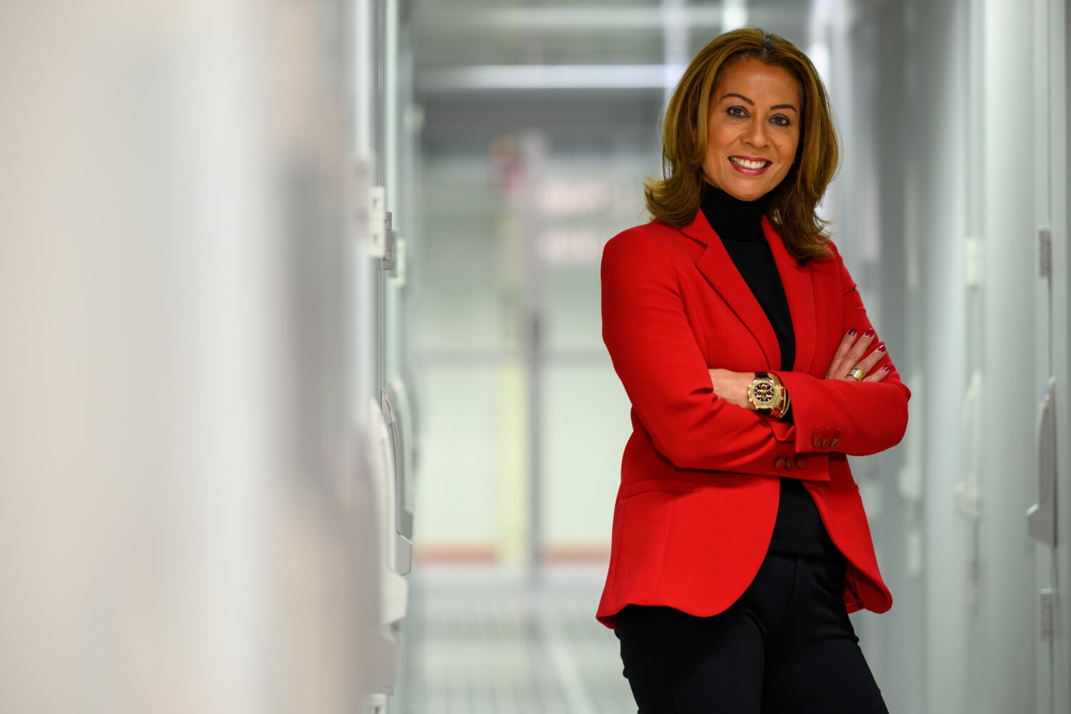 Een vrouw met een rode blazer en zwarte coltrui staat in een hal van NorthC met haar armen over elkaar en lacht naar de camera.