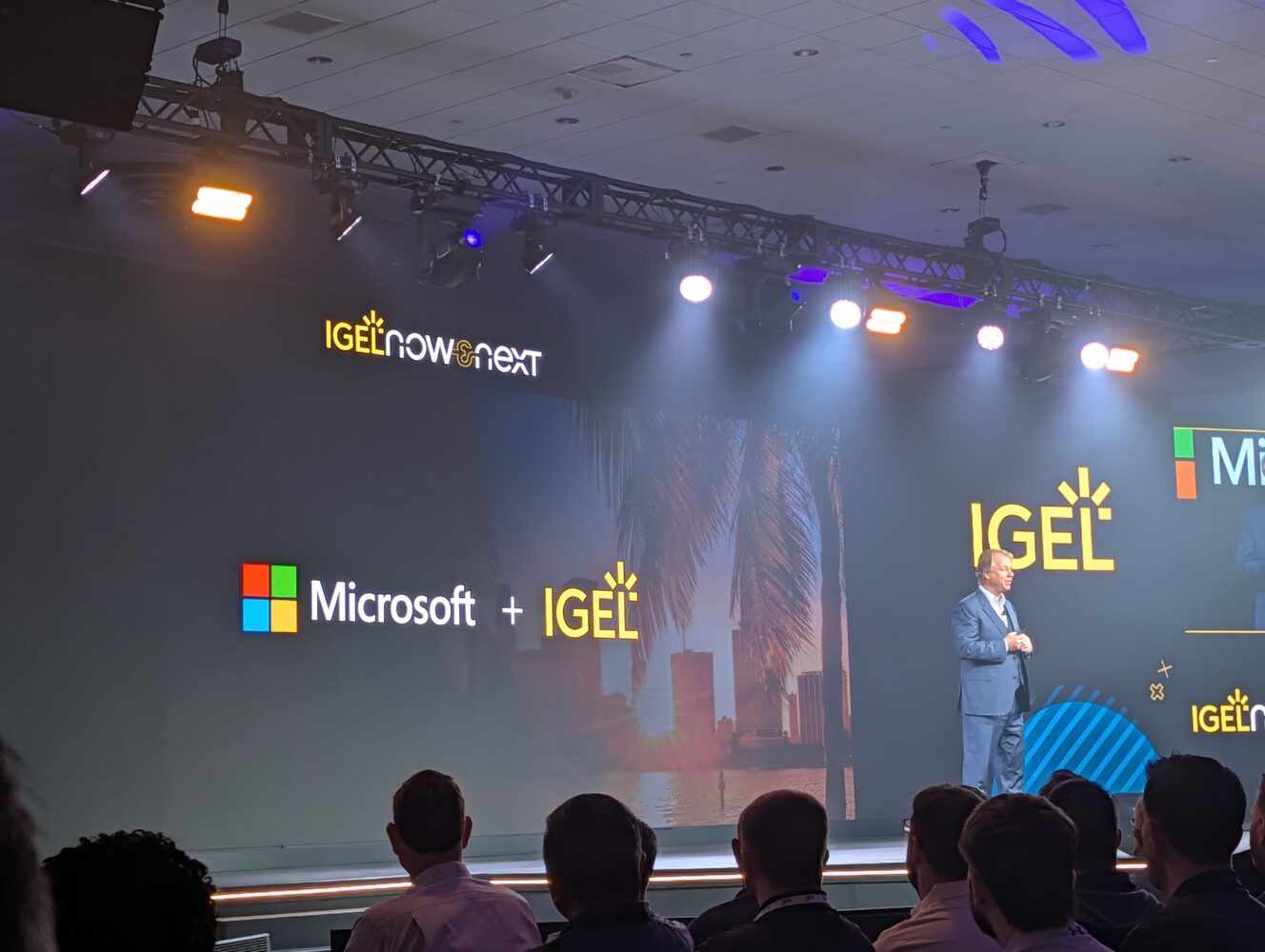 Een spreker staat op het podium tijdens een IGEL-evenement met op het scherm de logo's van Microsoft en IGEL en op de achtergrond een stadsgezicht.