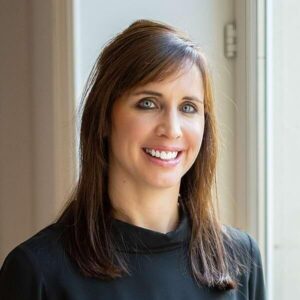 Vrouw met steil bruin haar en blauwe ogen draagt een zwart topje, glimlacht, staat binnen bij een raam met natuurlijk licht.