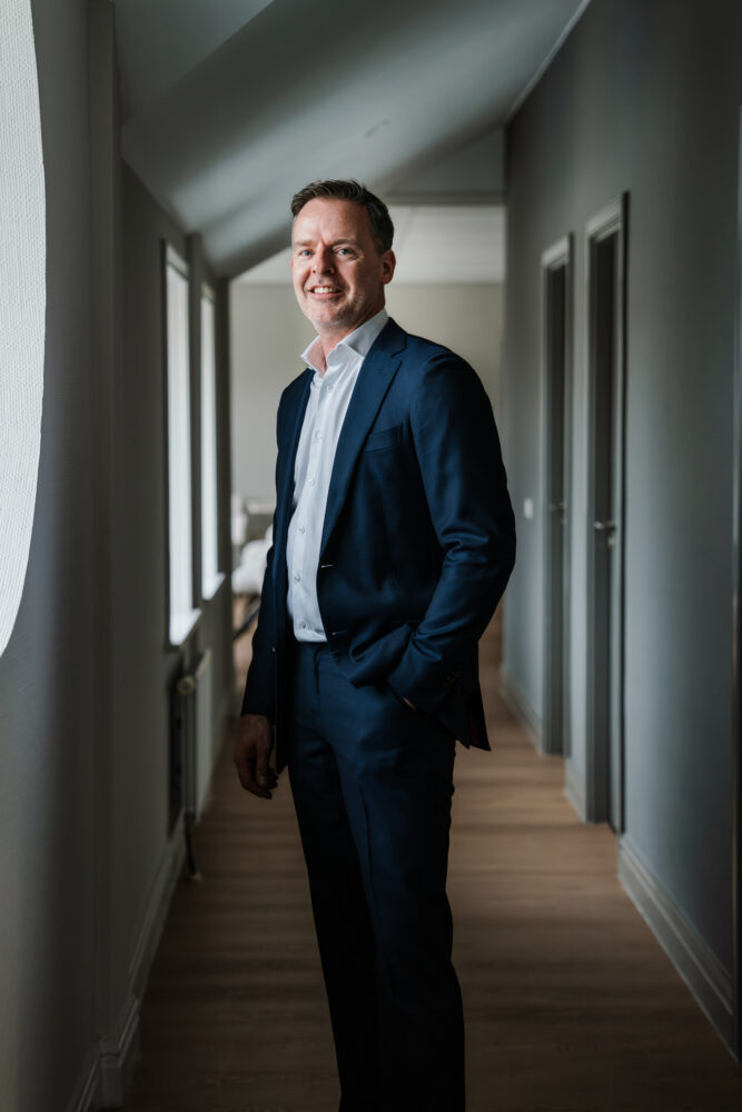 Een man in een marineblauw pak en wit overhemd staat in een gang met zijn hand in zijn zak en lacht naar de camera.