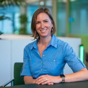 Een vrouw met een blauw overhemd zit aan een bureau en lacht naar de camera in een modern kantoor.