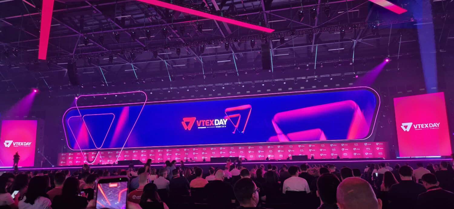 Een groot publiek zit tegenover een verlicht podium met VTEX DAY branding, agentic commerce graphics en VTEX AI Workspace displays op grote schermen in een ruim congrescentrum.