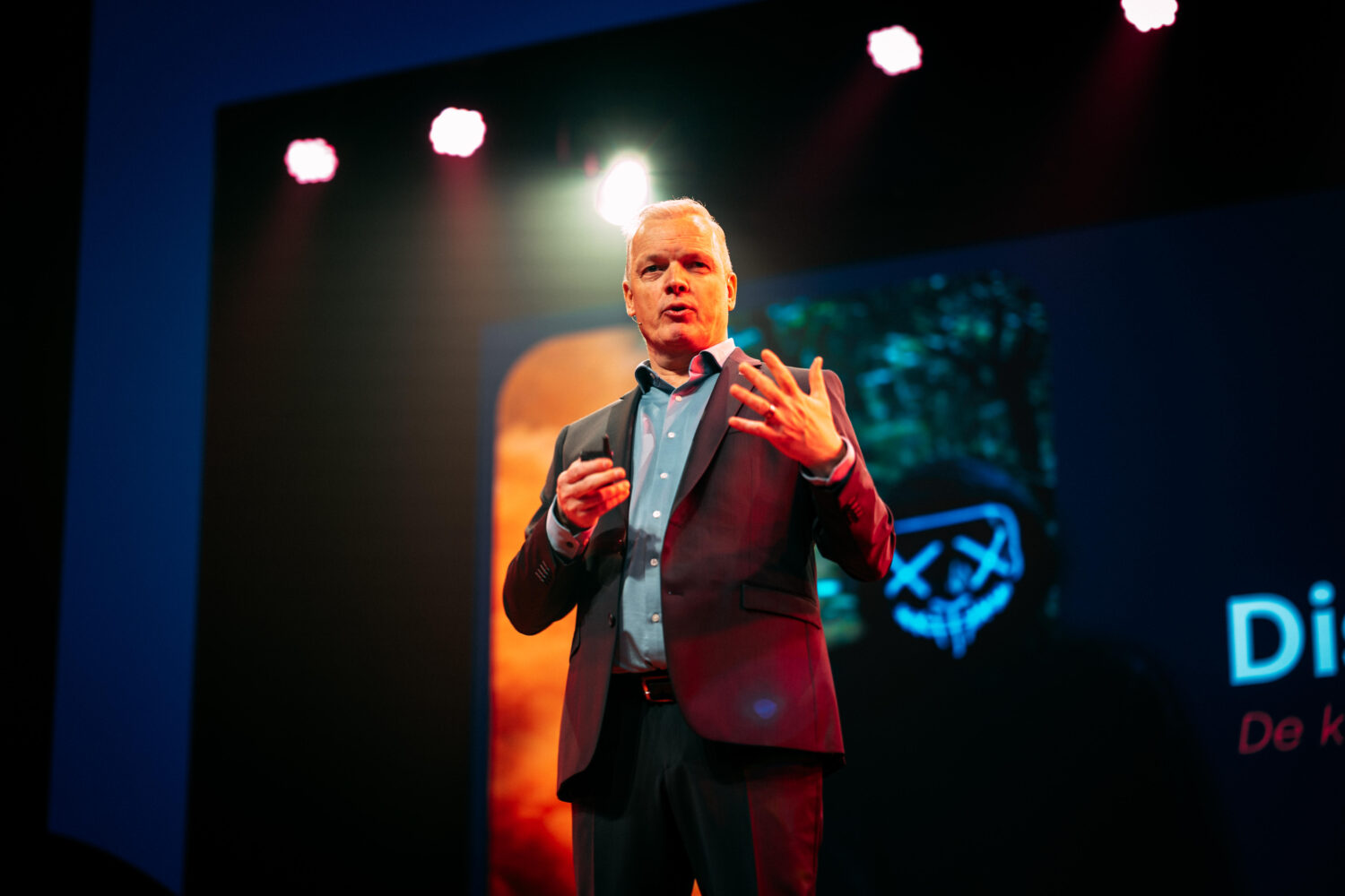 Een man in pak spreekt op het podium onder felle lampen, met achter hem een PQR-afbeelding van een gemaskerd gezicht met een kruis boven de ogen.
