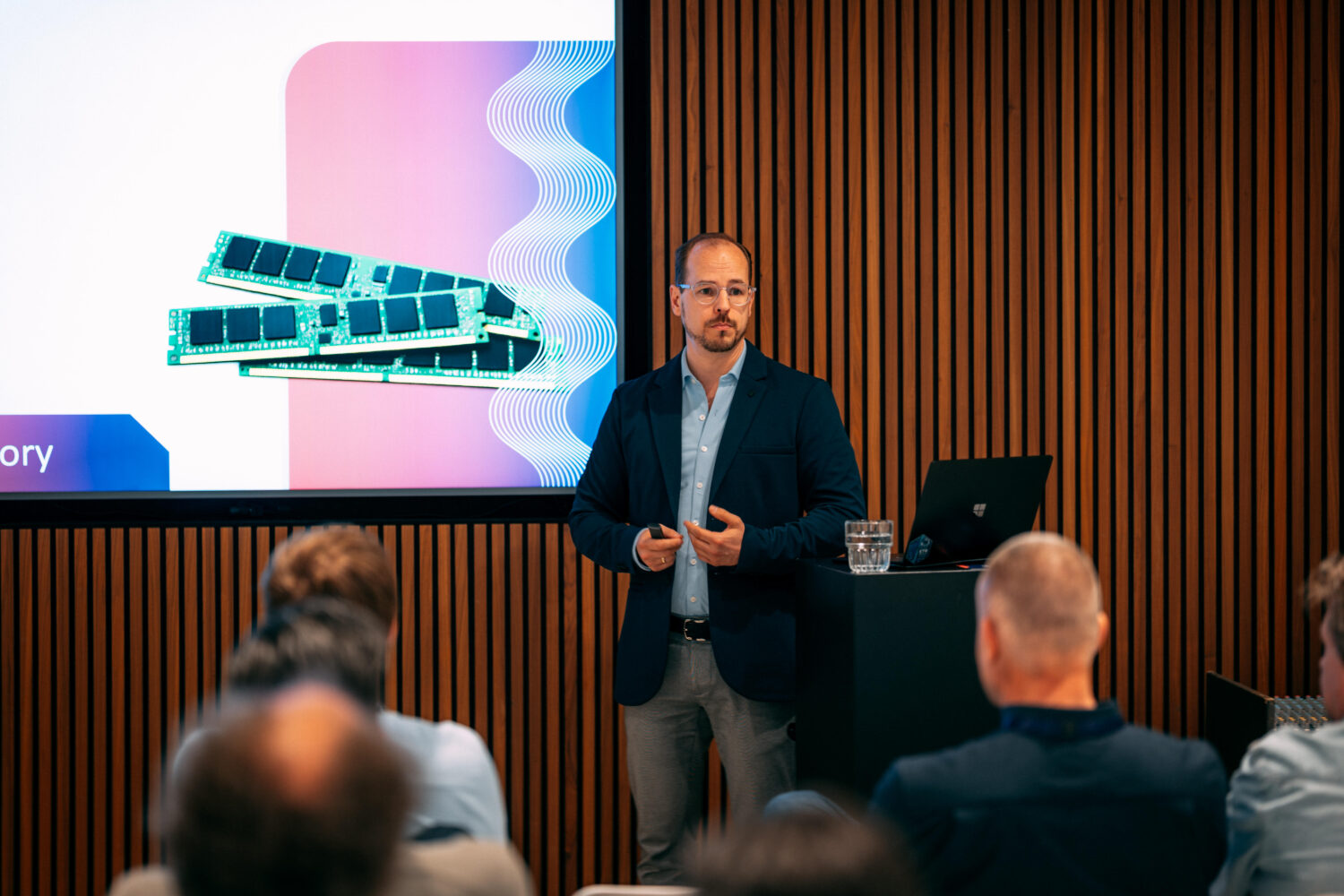 Een man in zakelijke kleding geeft een presentatie over PQR computergeheugenmodules aan een zittend publiek, met een dia van RAM-sticks op het scherm achter hem.