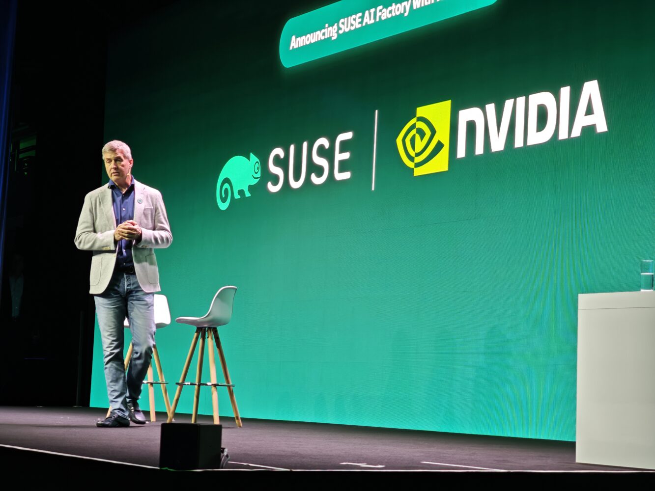 Een man staat op het podium tijdens SUSECON voor een groen scherm met de logo's van SUSE en NVIDIA, met een kruk en een tafeltje in de buurt, en benadrukt de rol van AI en soevereiniteit in het hedendaagse technologielandschap.
