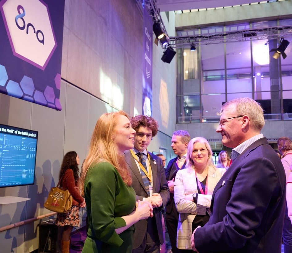Een groep mensen in zakelijke kleding praat met elkaar op een indoor evenement met digitale schermen en banners op de achtergrond.