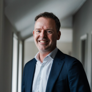 Een man in een donker pak en wit overhemd staat in een gang en lacht naar de camera.