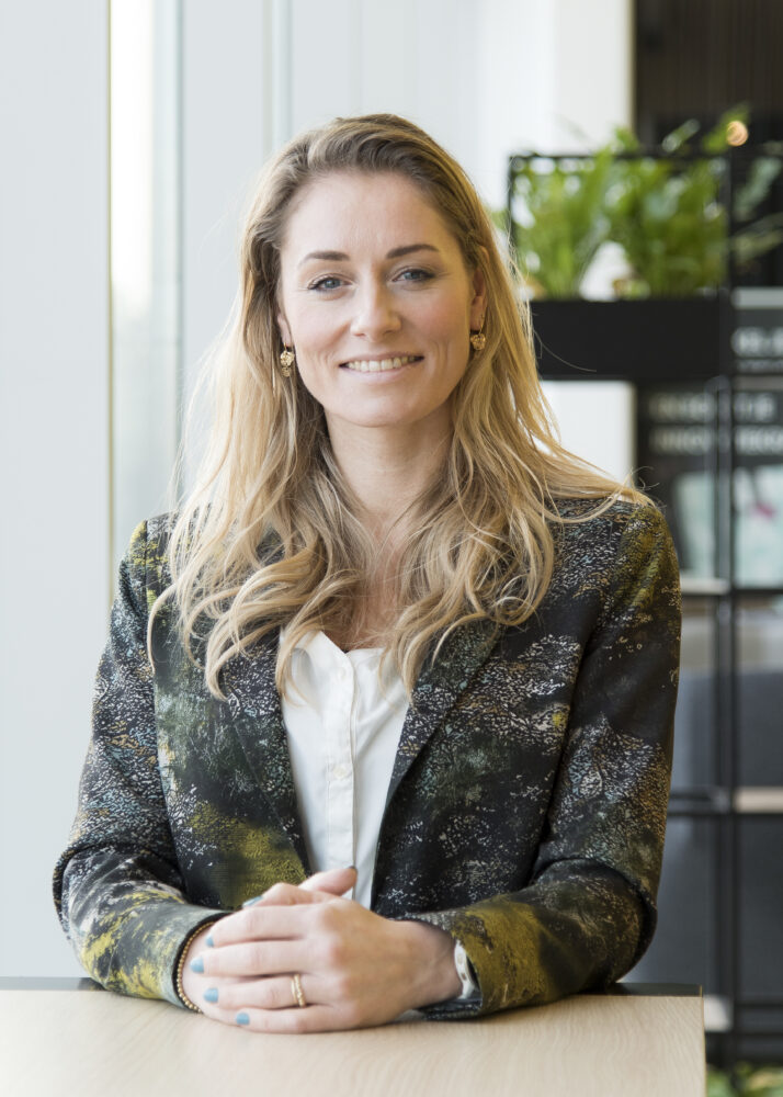 Een vrouw met lang blond haar zit aan een tafel, glimlacht, draagt een blazer met patroon en een witte blouse, met haar handen voor zich gevouwen. Op de achtergrond is een plant zichtbaar.