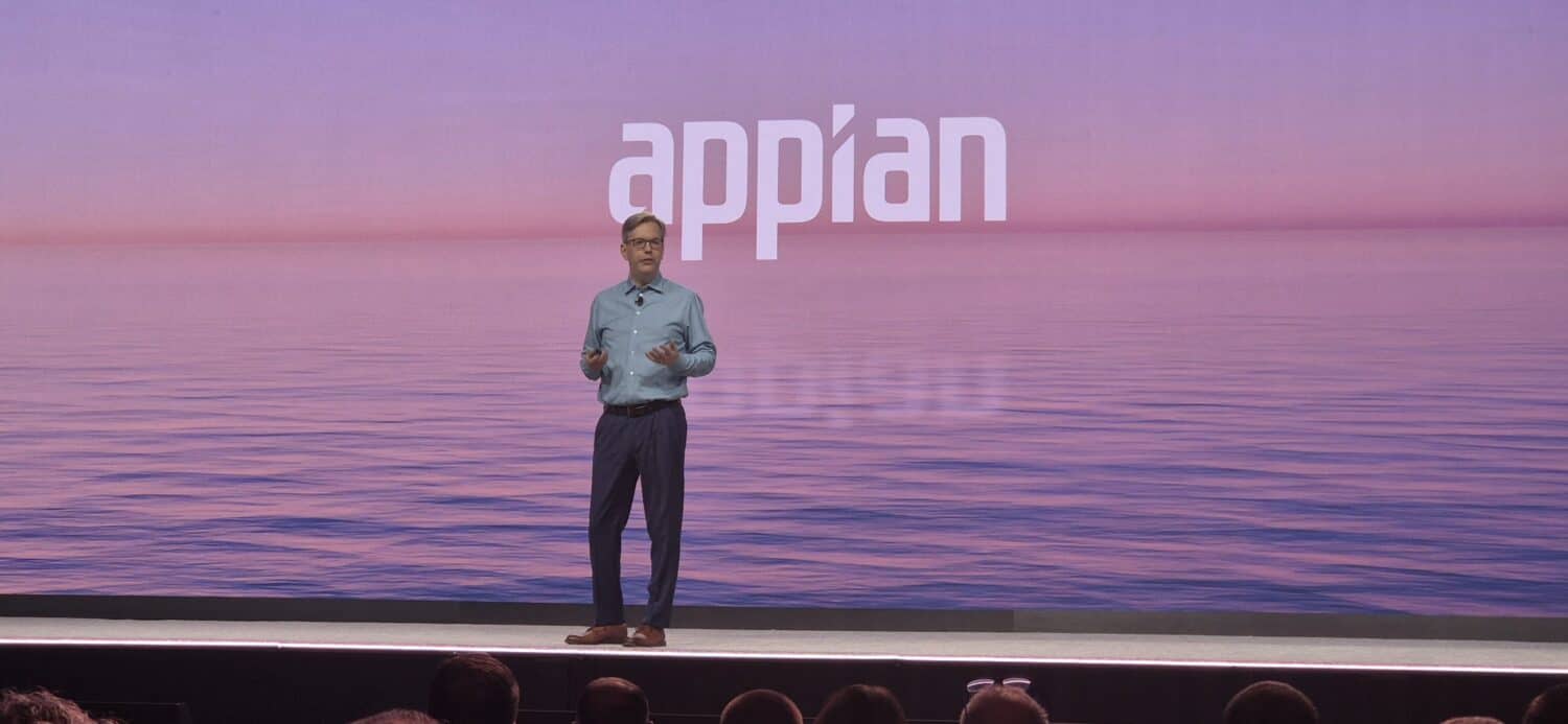 Een man staat op het podium en geeft een presentatie voor een scherm met het woord "Appian" en Appian AI procesautomatisering op een kalme zee achtergrond.
