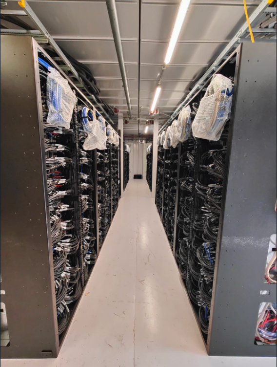 Een smal gangpad tussen rijen serverracks met bekabeling en apparatuur in een goed verlicht datacenter.