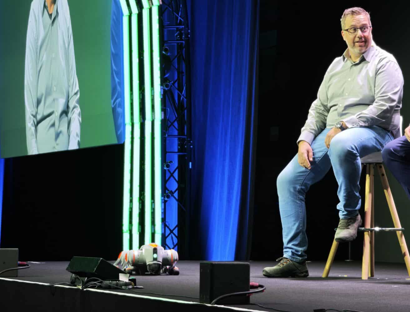 Een man in een lichtgrijs overhemd en spijkerbroek zit op een kruk op het podium, met een robot, elektronische apparatuur en ITQ AI-tools op de vloer, terwijl een groot scherm met een deel van zijn afbeelding op de achtergrond verschijnt.