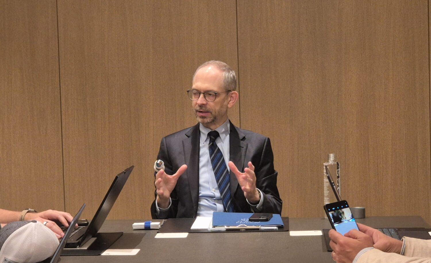 Een man in pak zit aan een tafel en bespreekt de procesautomatisering van Appian AI. Hij gebaart terwijl hij spreekt, met een laptop, map en waterfles voor zich. Twee andere mensen zijn gedeeltelijk zichtbaar.