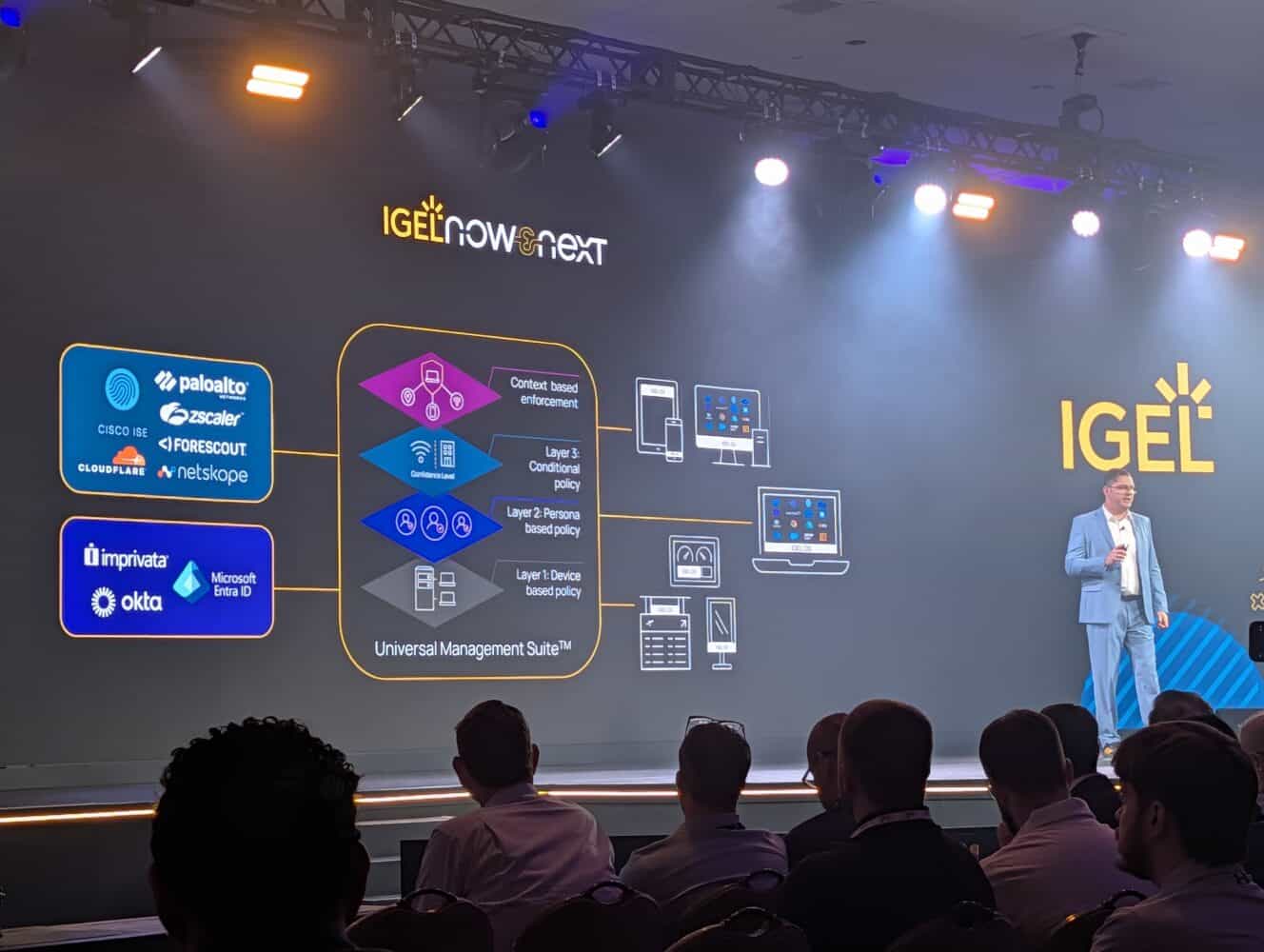 Een spreker presenteert op het podium tijdens het IGEL-evenement, staand naast een groot scherm waarop oplossingen voor cyberbeveiliging en -beheer van verschillende technologiebedrijven worden getoond.