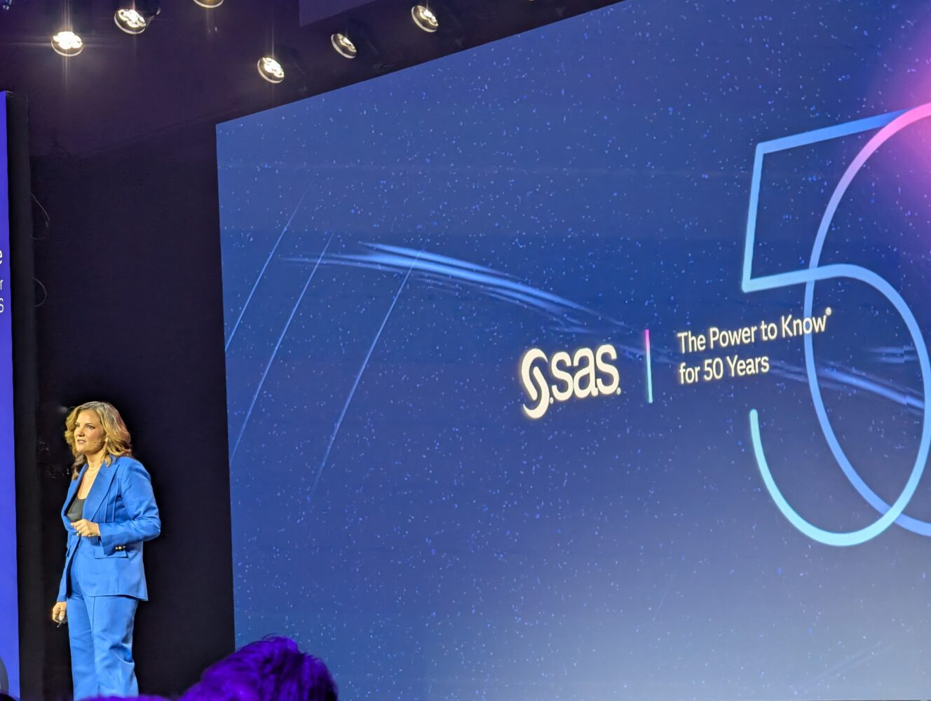 Een persoon in een blauw pak staat op het podium naast een groot scherm met het SAS-logo en de tekst "The Power to Know for 50 Years", waarmee de innovatie-erfenis van SAS wordt benadrukt.
