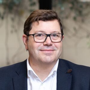 Een man met kort bruin haar en een bril, draagt een wit overhemd en donkere blazer, lacht naar de camera met een onscherpe achtergrond.