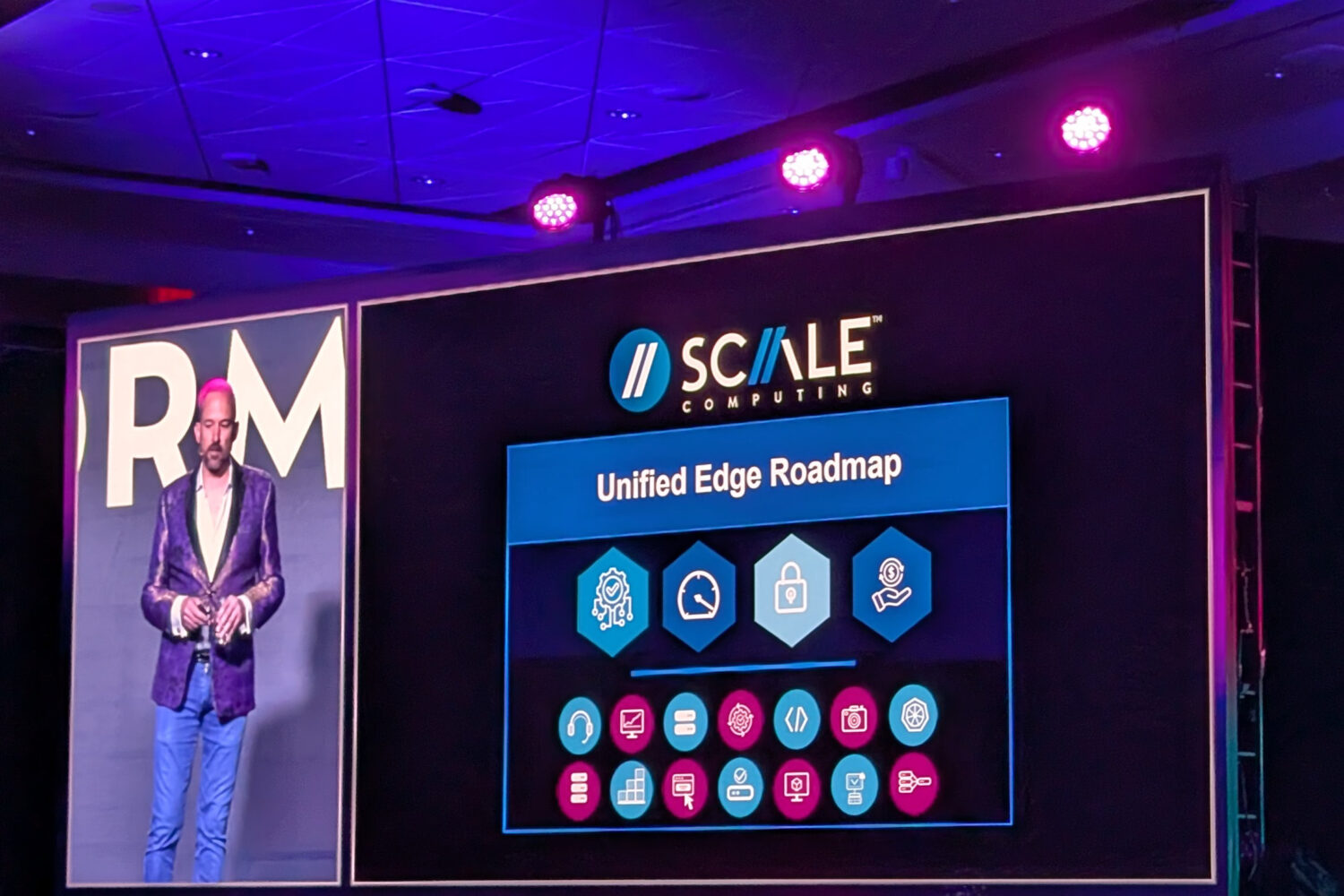 Scale_Unified_Edge Een presentator staat naast een groot scherm met daarop de "SCALE Computing Unified Edge Roadmap" met pictogrammen en roadmap-elementen onder podiumverlichting.