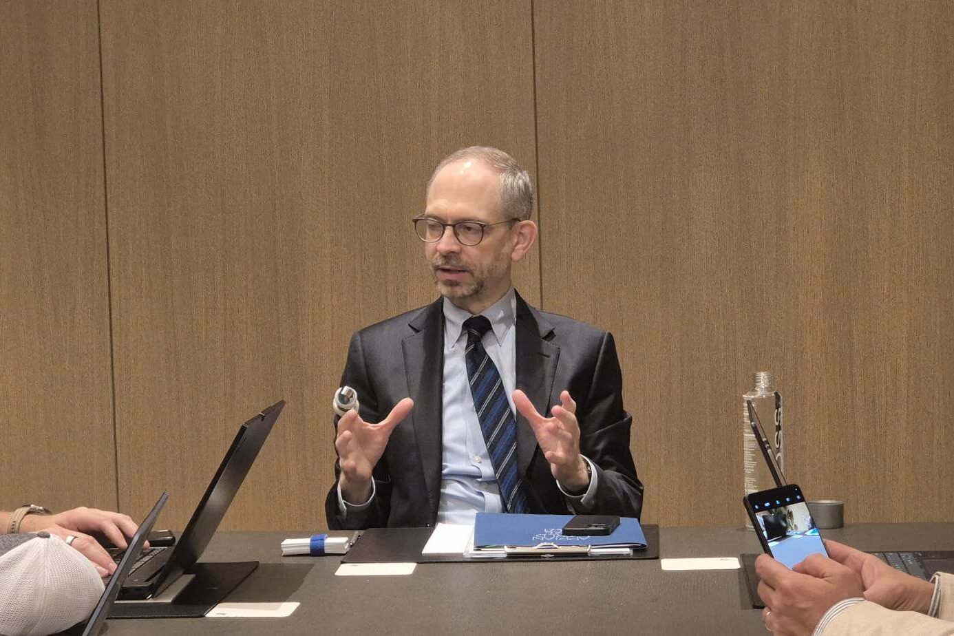 Een man in pak spreekt terwijl hij aan een tafel zit met twee andere mannen. De ene gebruikt een laptop en de andere houdt een telefoon vast. De achtergrond is een houten muur.