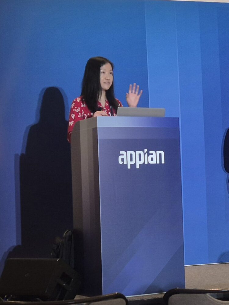 Een vrouw in een rood gebloemd shirt staat op een podium met de tekst "Appian" en spreekt over Appian AI procesautomatisering en gebaart met haar rechterhand tijdens een conferentie.