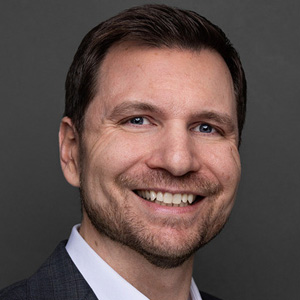 Een man met kort bruin haar en een getrimde baard glimlacht naar de camera, draagt een donker colbert en wit overhemd tegen een effen donkere achtergrond.